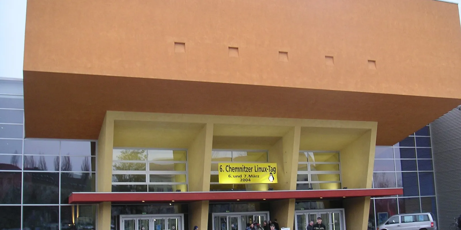 Three entrance doors in glass front framed by a yellow concrete construction, crowned by an immense orange colored auditorium block