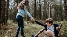 Boy crouching o a fallen tree, girl lending him a hand to get back on his feet