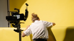 Woman turning her back to a camera in the foreground, leaning over a yellow wall with a higher yellow wall in the background