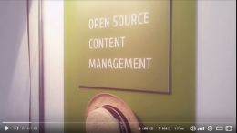 Video-Standbild: Schild an einer Messestand-Rückwand mit englischem Text Open Source Content Management, darunter hängt ein Strohhut
