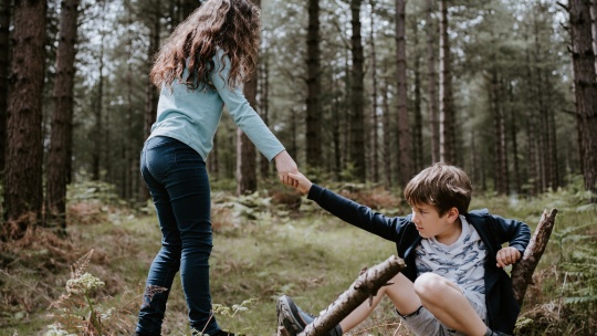 Ein Junge hockt auf einem liegenden Baumstamm, ein Mädchen reicht ihm die Hand, um ihm aufzuhelfen