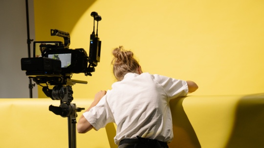 Woman turning her back to a camera in the foreground, leaning over a yellow wall with a higher yellow wall in the background