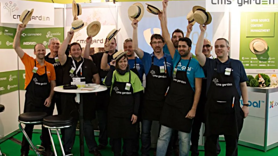 11 Menschen mit schwarzen Gartenschürzen an einem Messestand schwenken ihre Strohhüte